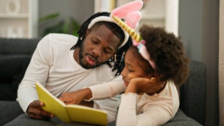 Foto de Afro-americanos padre e hija usando divertido diadema acostado en sofá lectura libro en casa - Imagen libre de derechos