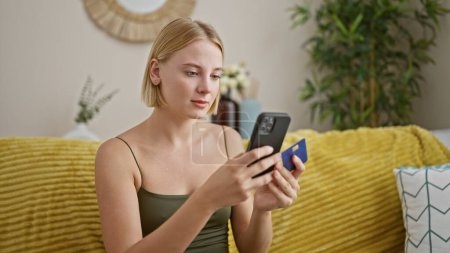 Photo for Young blonde woman shopping with smartphone and credit card sitting on sofa at home - Royalty Free Image
