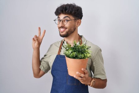 Téléchargez les photos : Homme arabe avec barbe tenant pot de plante verte souriant en regardant la caméra montrant les doigts faisant signe de victoire. numéro deux. - en image libre de droit