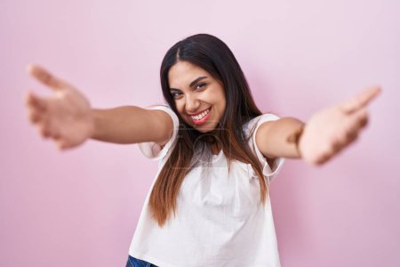 Photo for Young arab woman standing over pink background looking at the camera smiling with open arms for hug. cheerful expression embracing happiness. - Royalty Free Image