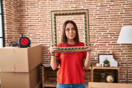 Foto de Mujer hispana joven en casa nueva sosteniendo el marco vacío sonriendo mirando hacia un lado y mirando hacia otro lado pensando. - Imagen libre de derechos