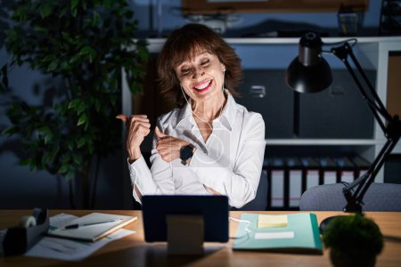 Foto de Mujer de mediana edad que trabaja en la oficina por la noche señalando hacia atrás con la mano y los pulgares hacia arriba, sonriendo confiada - Imagen libre de derechos