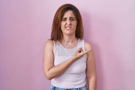 Photo for Brunette woman standing over pink background pointing aside worried and nervous with forefinger, concerned and surprised expression - Royalty Free Image