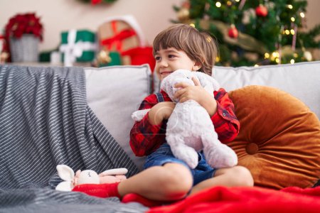 Foto de Adorable niño abrazando conejo muñeca sentado en sofá por árbol de Navidad en casa - Imagen libre de derechos