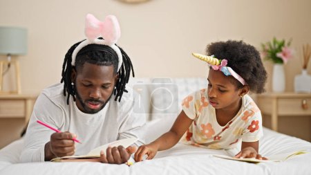 Foto de Afroamericanos padre e hija usando divertido dibujo de la diadema en el cuaderno acostado en la cama en el dormitorio - Imagen libre de derechos