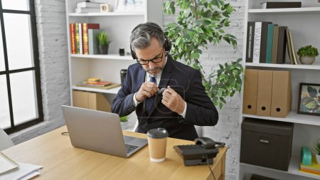 Photo for Attractive grey-haired young hispanic man mastering the office call center game, video chatting with client on laptop, adorned in suit and tie, listening to cool tunes. - Royalty Free Image