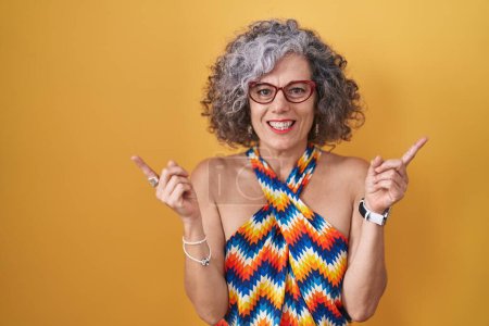 Foto de Mujer de mediana edad con el pelo gris de pie sobre el fondo amarillo sonriendo confiado señalando con los dedos a diferentes direcciones. espacio de copia para publicidad - Imagen libre de derechos