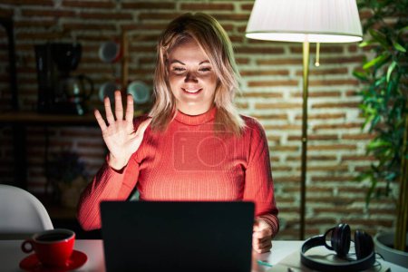 Photo for Blonde woman using laptop at night at home showing and pointing up with fingers number five while smiling confident and happy. - Royalty Free Image
