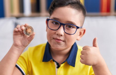 Foto de Joven niño hispano sosteniendo moneda criptomoneda tron sonriendo feliz y positivo, pulgar hacia arriba haciendo excelente y signo de aprobación - Imagen libre de derechos