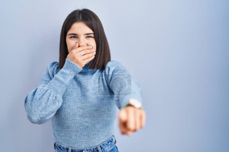 Téléchargez les photos : Young hispanic woman standing over blue background laughing at you, pointing finger to the camera with hand over mouth, shame expression - en image libre de droit