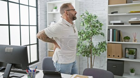 Photo for Unhappy young man worker suffering from backache while standing indoors at the office, serious business professional touching injured spinal column, worried about painful back injury at workplace - Royalty Free Image