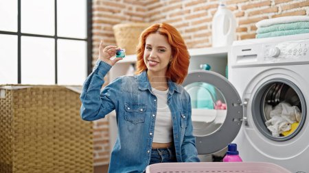 Photo for Young redhead woman washing clothes holding detergent bag at laundry room - Royalty Free Image