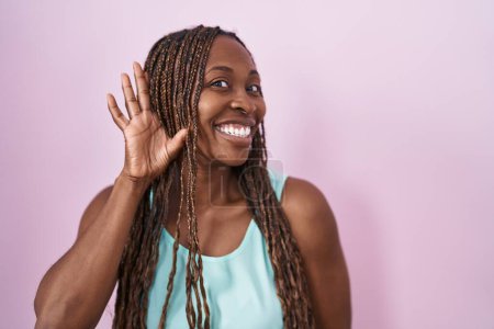 Téléchargez les photos : Femme afro-américaine debout sur fond rose souriant avec la main sur l'oreille en écoutant une rumeur ou des commérages. concept de surdité. - en image libre de droit