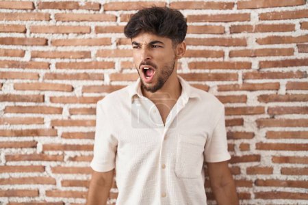 Téléchargez les photos : Homme arabe avec barbe debout sur fond de mur de briques en colère et fou hurlant frustré et furieux, criant de colère. rage et concept agressif. - en image libre de droit