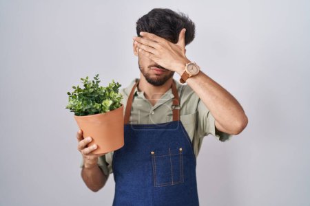 Téléchargez les photos : Homme arabe avec barbe tenant pot de plante verte couvrant les yeux avec la main, l'air grave et triste. concept de non-visibilité, de dissimulation et de rejet - en image libre de droit