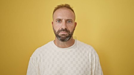 Photo for Handsome young man, with a concentrated expression, nonchalantly standing against an isolated yellow background, exudes coolness while looking right into the camera. - Royalty Free Image