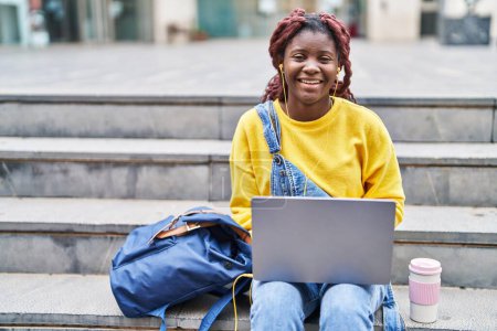 Téléchargez les photos : Étudiante afro-américaine utilisant un ordinateur portable et des écouteurs assis sur les escaliers à l'université - en image libre de droit