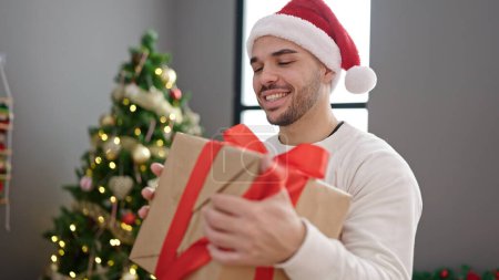 Foto de Joven hombre hispano sonriendo confiado sosteniendo regalo de Navidad en casa - Imagen libre de derechos