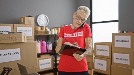 Téléchargez les photos : Femme âgée aux cheveux gris parlant bénévolement sur smartphone prenant des notes au centre de bienfaisance - en image libre de droit