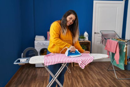 Photo for Young beautiful plus size woman smiling confident ironing clothes at laundry room - Royalty Free Image