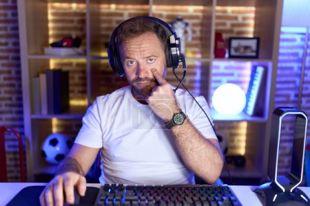 Téléchargez les photos : Homme d'âge moyen avec barbe jouant à des jeux vidéo portant un casque pointant vers l'œil vous regardant geste, expression suspecte - en image libre de droit