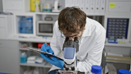 Photo for Serious young man scientist, a bearded blond, meticulously taking notes at lab table, engrossed in medical research under microscope at a bustling science laboratory. - Royalty Free Image