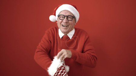 Téléchargez les photos : Senior portant un chapeau de Noël regardant à l'intérieur de la chaussette sur fond rouge isolé - en image libre de droit