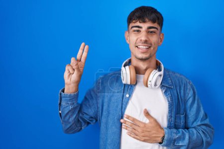 Téléchargez les photos : Jeune homme hispanique debout sur fond bleu souriant jurer avec la main sur la poitrine et les doigts vers le haut, faisant un serment de fidélité promesse - en image libre de droit