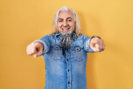 Téléchargez les photos : Homme d'âge moyen avec les cheveux gris debout sur fond jaune pointant vers vous et la caméra avec les doigts, souriant positif et joyeux - en image libre de droit