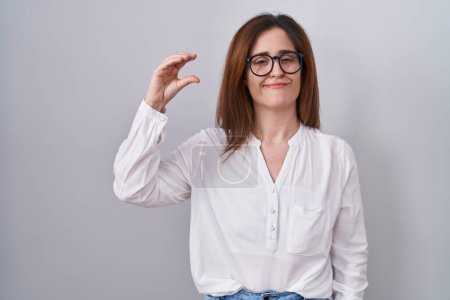 Téléchargez les photos : Femme brune debout sur fond blanc isolé geste souriant et confiant avec la main faisant signe de petite taille avec les doigts regardant et la caméra. concept de mesure. - en image libre de droit