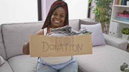 Foto de Hermosa mujer afroamericana sonriendo mientras prepara un paquete de ropa para la donación en casa - Imagen libre de derechos