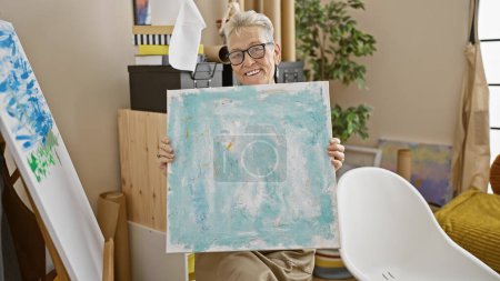 Photo for Confident, grey-haired senior woman artist, smiling and holding her artistic draw, revels in the joy of creativity in a bustling art studio - Royalty Free Image