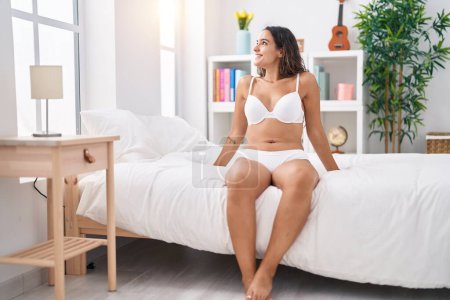 Foto de Joven mujer hispana hermosa usando lencería sentada en la cama en el dormitorio - Imagen libre de derechos