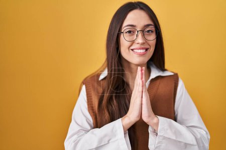 Téléchargez les photos : Jeune femme brune debout sur fond jaune portant des lunettes priant avec les mains ensemble demandant pardon en souriant confiant. - en image libre de droit