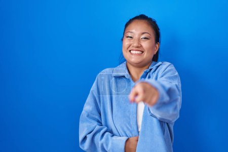 Foto de Asiática joven mujer de pie sobre fondo azul riéndose de usted, señalando el dedo a la cámara con la mano sobre el cuerpo, vergüenza expresión - Imagen libre de derechos