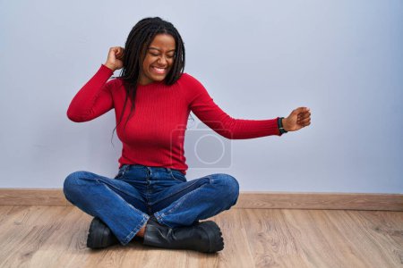 Téléchargez les photos : Jeune afro-américain avec des tresses assises sur le sol à la maison dansant heureux et joyeux, souriant décontracté et confiant écoutant de la musique - en image libre de droit