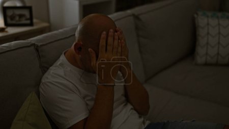 Téléchargez les photos : Jeune homme hispanique stressé assis sur le canapé portant des écouteurs à la maison - en image libre de droit