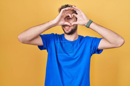 Photo for Hispanic man with beard standing over yellow background doing heart shape with hand and fingers smiling looking through sign - Royalty Free Image