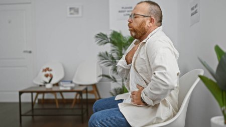 Handsome middle-aged caucasian man, with beard and glasses, sitting unhappily in chair, worriedly coughing in waiting room amid covid19 fears. an upset portrait of indoor illness stress.