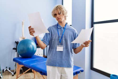Foto de Joven pisioterapeuta hombre rubio leyendo documento de pie en la clínica de rehabilitación - Imagen libre de derechos