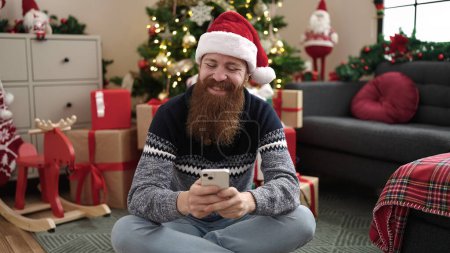 Téléchargez les photos : Jeune rousse homme en utilisant smartphone assis sur le sol par arbre de Noël à la maison - en image libre de droit