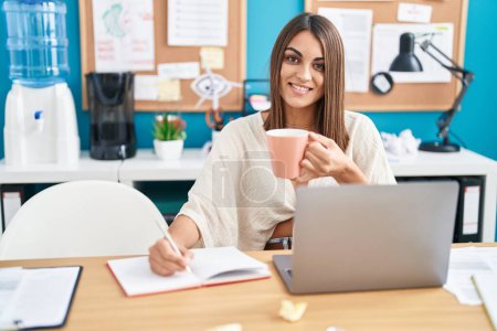 Photo for Young beautiful hispanic woman business worker writing on notebook drinking coffee at office - Royalty Free Image