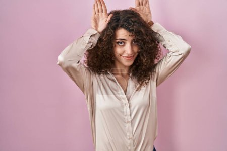 Foto de Hispanic woman with curly hair standing over pink background doing bunny ears gesture with hands palms looking cynical and skeptical. easter rabbit concept. - Imagen libre de derechos