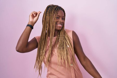 Téléchargez les photos : Femme afro-américaine avec des cheveux tressés debout sur fond rose dansant heureux et joyeux, souriant décontracté et confiant écoutant de la musique - en image libre de droit