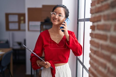 Foto de Young chinese woman business worker talking on smartphone holding clipboard at office - Imagen libre de derechos