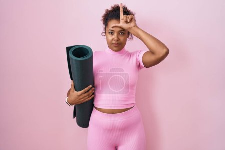 Photo for Young hispanic woman with curly hair holding yoga mat over pink background making fun of people with fingers on forehead doing loser gesture mocking and insulting. - Royalty Free Image