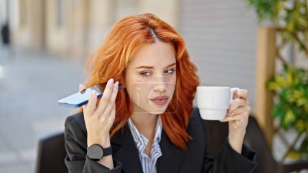 Téléchargez les photos : Jeune rousse femme d'affaires ouïe message vocal par smartphone boire du café à la terrasse du café - en image libre de droit