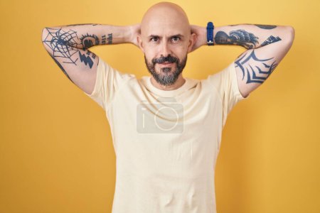 Foto de Hombre hispano con tatuajes de pie sobre fondo amarillo relajante y estiramiento, brazos y manos detrás de la cabeza y el cuello sonriendo feliz - Imagen libre de derechos