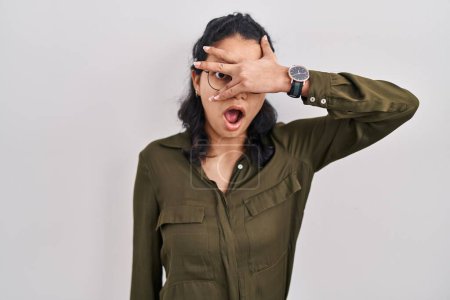 Photo for Hispanic woman with dark hair standing over isolated background peeking in shock covering face and eyes with hand, looking through fingers with embarrassed expression. - Royalty Free Image