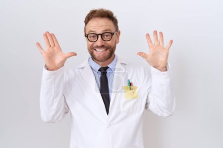 Foto de Médico de mediana edad hombre con barba con abrigo blanco mostrando y apuntando hacia arriba con los dedos número diez mientras sonríe confiado y feliz. - Imagen libre de derechos